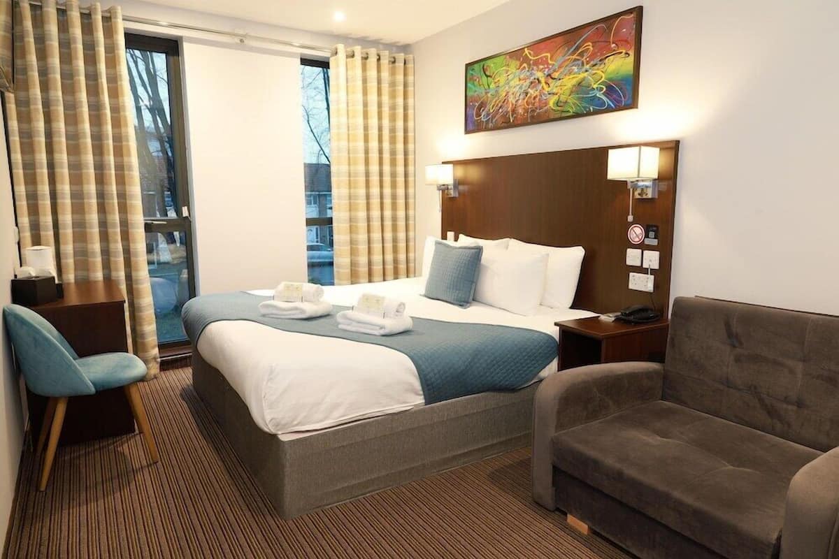 Une chambre d’hôtel avec un lit, un bureau, une chaise, un canapé, un tableau et une fenêtre avec des rideaux.