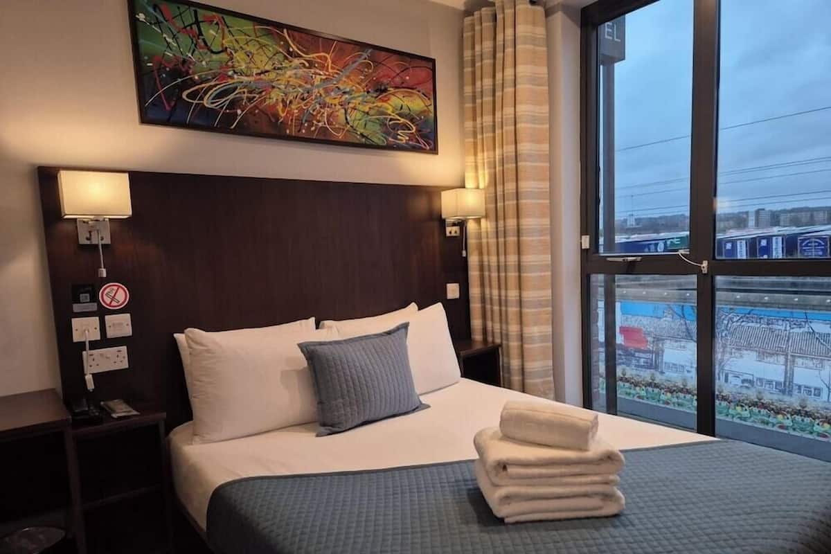 Une chambre d’hôtel avec un grand lit, une tête de lit, une table de chevet, une lampe fixée au mur, un tableau et une vue sur le paysage urbain par la fenêtre.