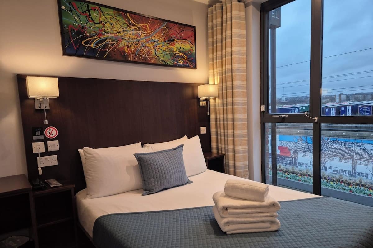 Une chambre d’hôtel avec un grand lit, une tête de lit, une table de chevet, une lampe fixée au mur, un tableau et une fenêtre donnant sur des immeubles.