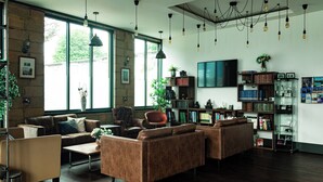 Ein geräumiges Wohnzimmer mit Ledersofas, einem Couchtisch und einem Bücherregal voller Bücher und Dekoration. Große Fenster lassen viel natürliches Licht herein, und ein Flachbildfernseher ist an der Wand befestigt.