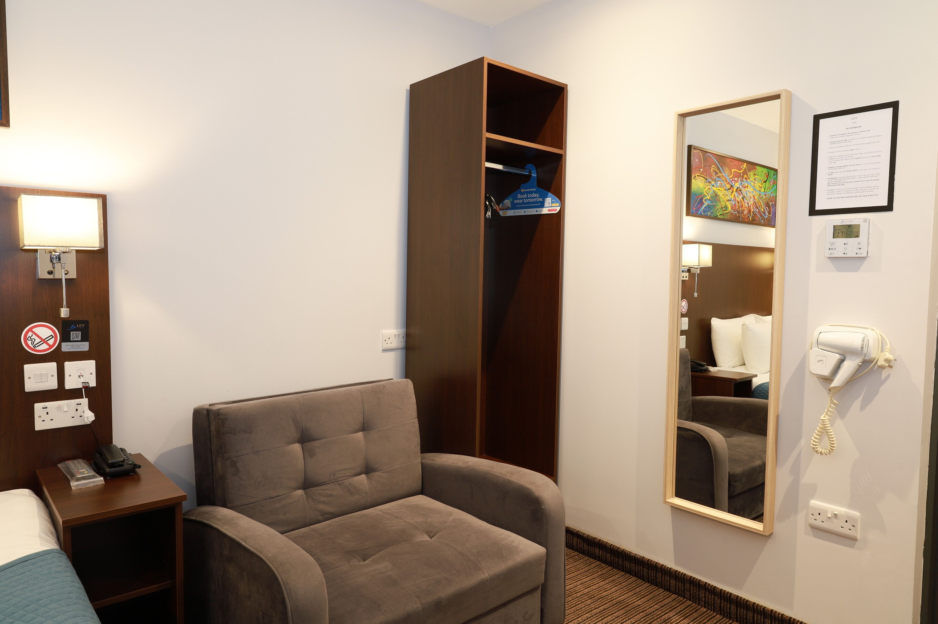 A hotel room with a single bed, a sofa, a wardrobe, a mirror, a hairdryer, and a wall-mounted phone.