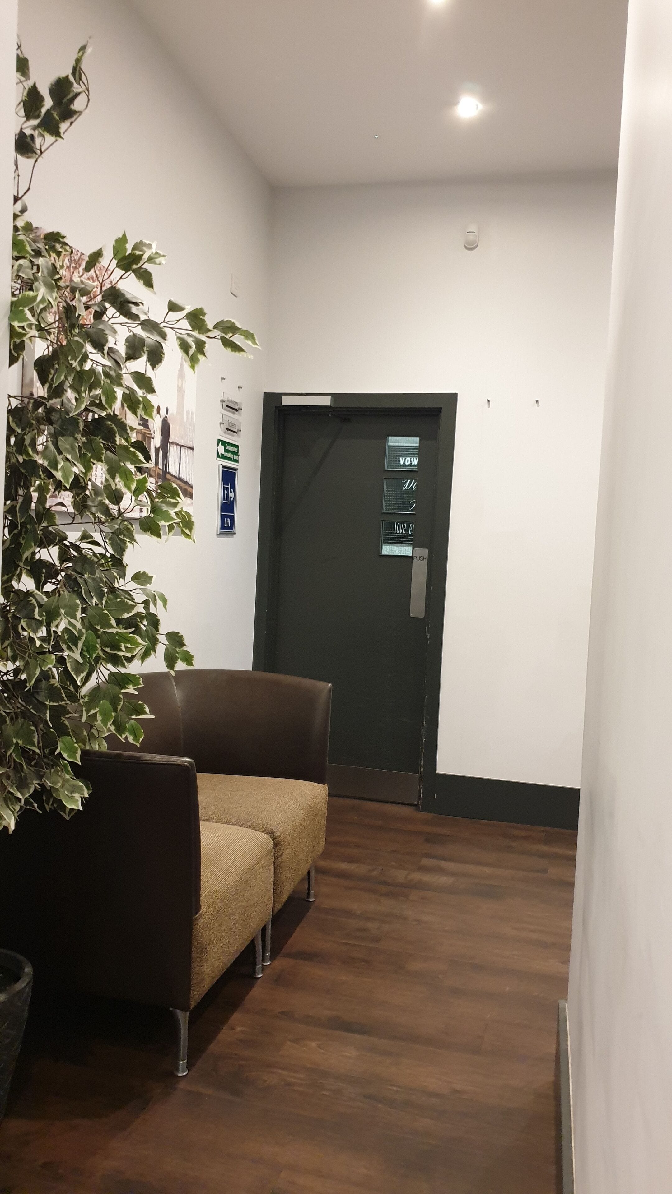 Un pasillo con piso marrón oscuro, un sofá marrón y una planta en maceta. Hay una puerta con un letrero que dice 'VOW' y otro que indica 'PUSH'.