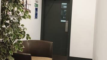 Un pasillo con piso marrón oscuro, un sofá marrón y una planta en maceta. Hay una puerta con un letrero que dice 'VOW' y otro que indica 'PUSH'.