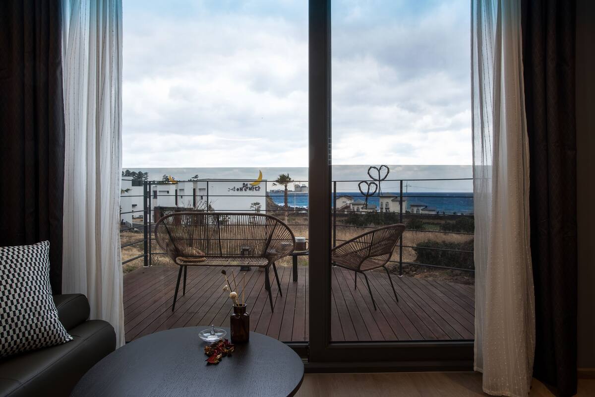 A balcony with a view of the sea, furnished with a sofa and chairs, and a table with a vase of flowers.