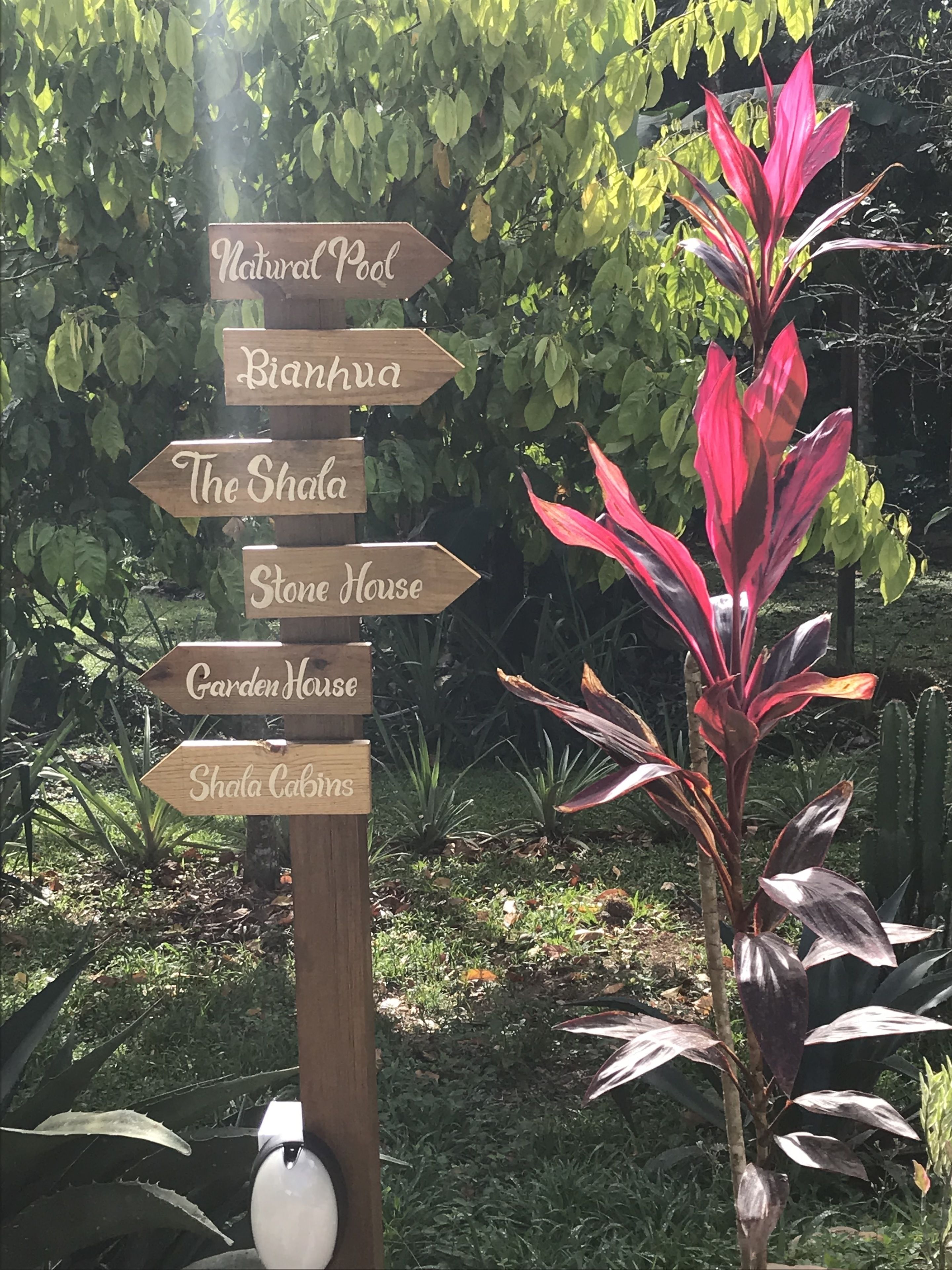 Un cartel de madera con flechas direccionales que indican varios lugares como 'Natural Pool', 'Bianhua', 'The Shala', 'Stone House', 'Garden House' y 'Shala Cabins'.