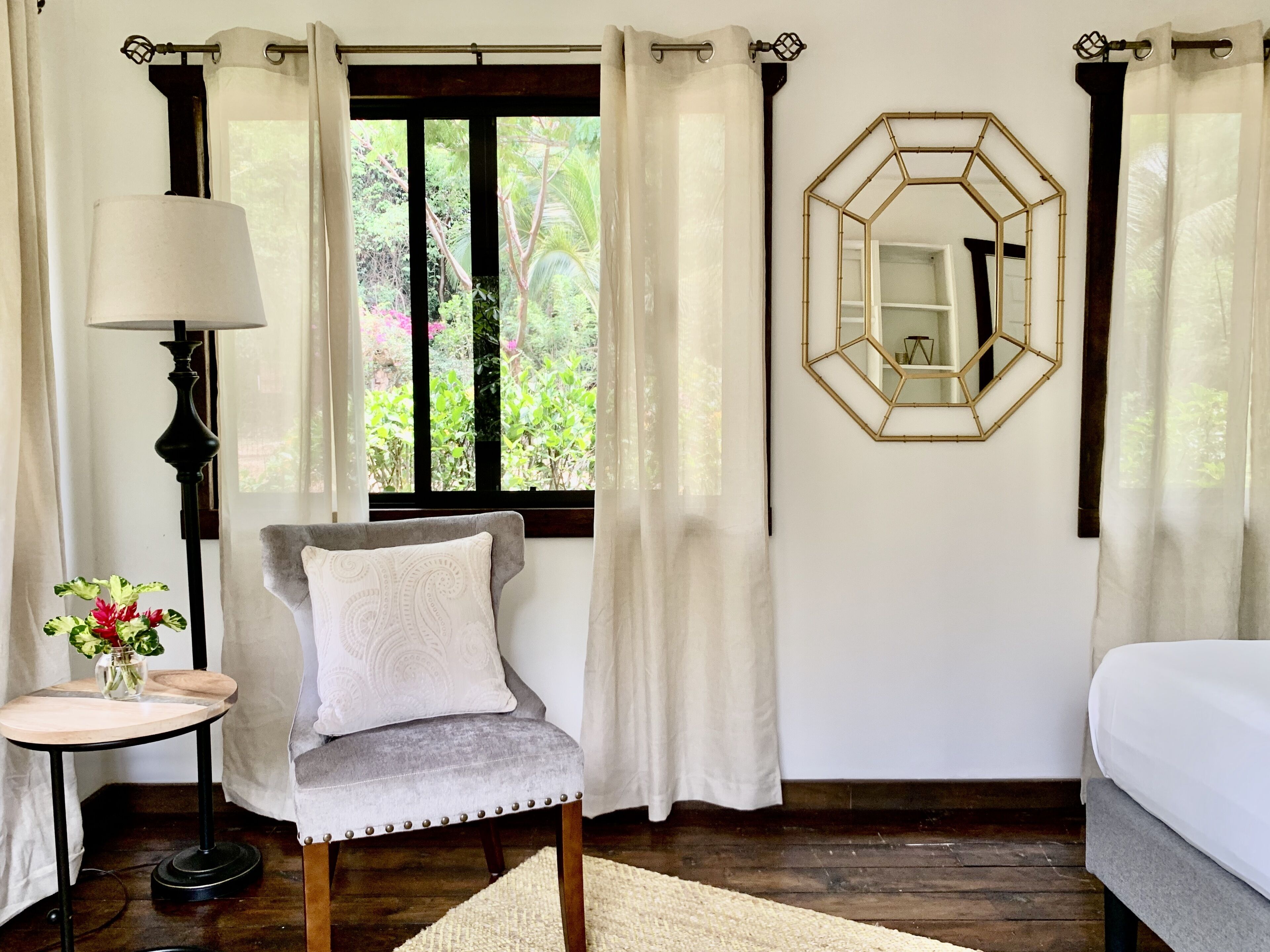 Una habitación con un sillón gris, una mesita con un florero, una lámpara y un ventanal con cortinas.