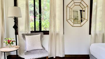 Una habitación con un sillón gris, una mesita con un florero, una lámpara y un ventanal con cortinas.