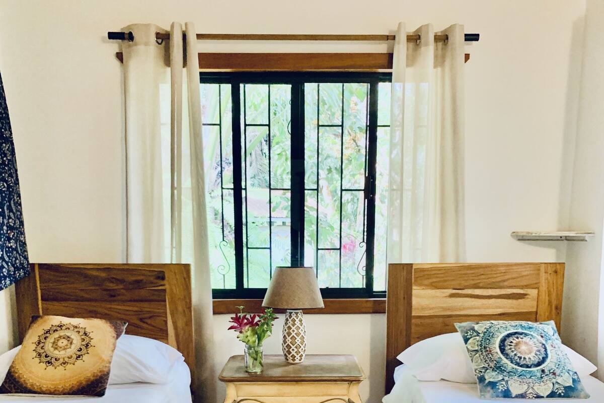 Ein Zimmer mit zwei Betten, einem kleinen Tisch mit Lampe und einem Fenster mit Vorhängen.