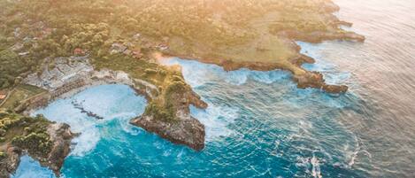 Vista aérea de una zona costera con mar azul cristalino, formaciones rocosas y un pequeño asentamiento con edificios y vegetación.