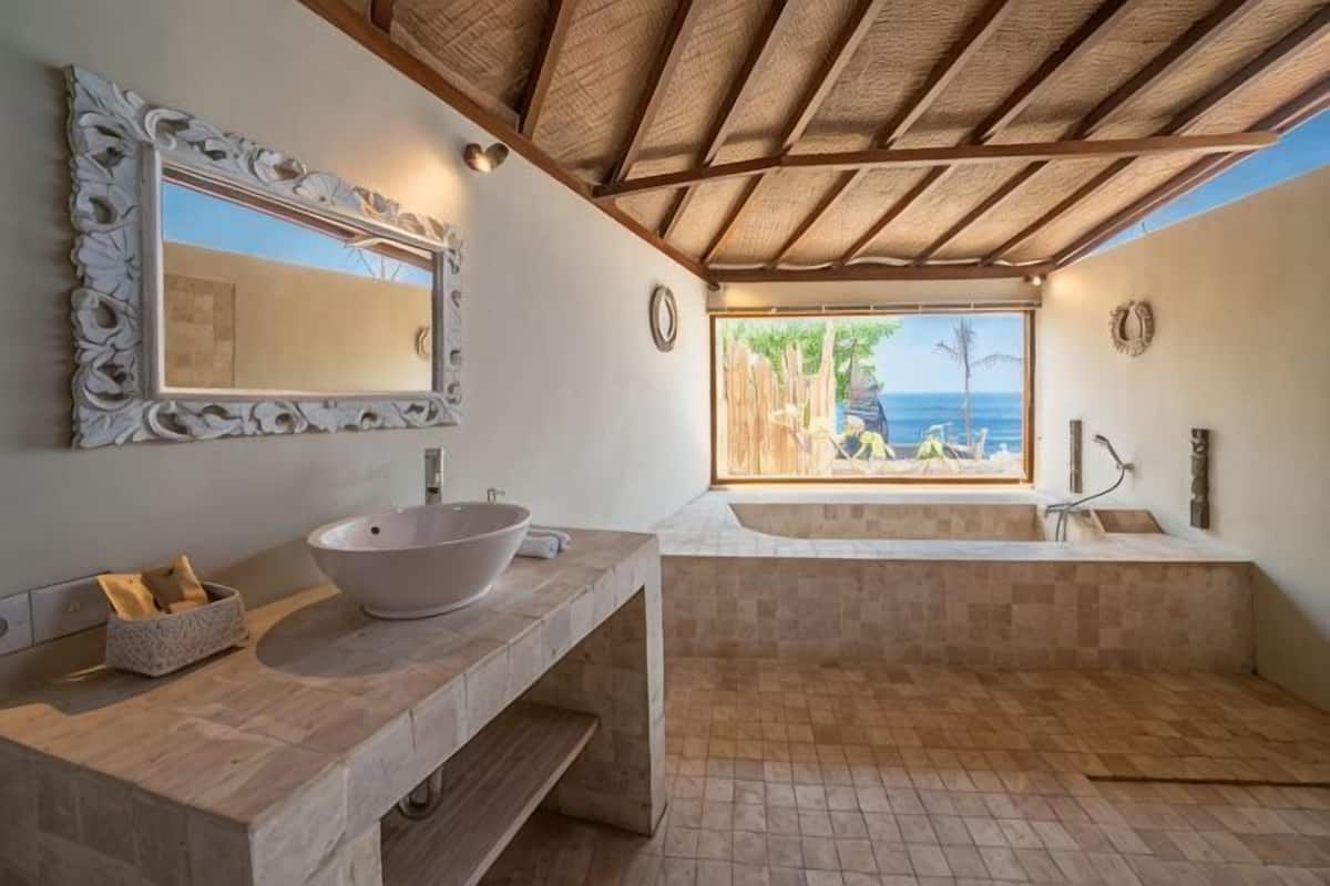 A bathroom with a large mirror, a bowl sink, a bathtub, and a view of the beach.