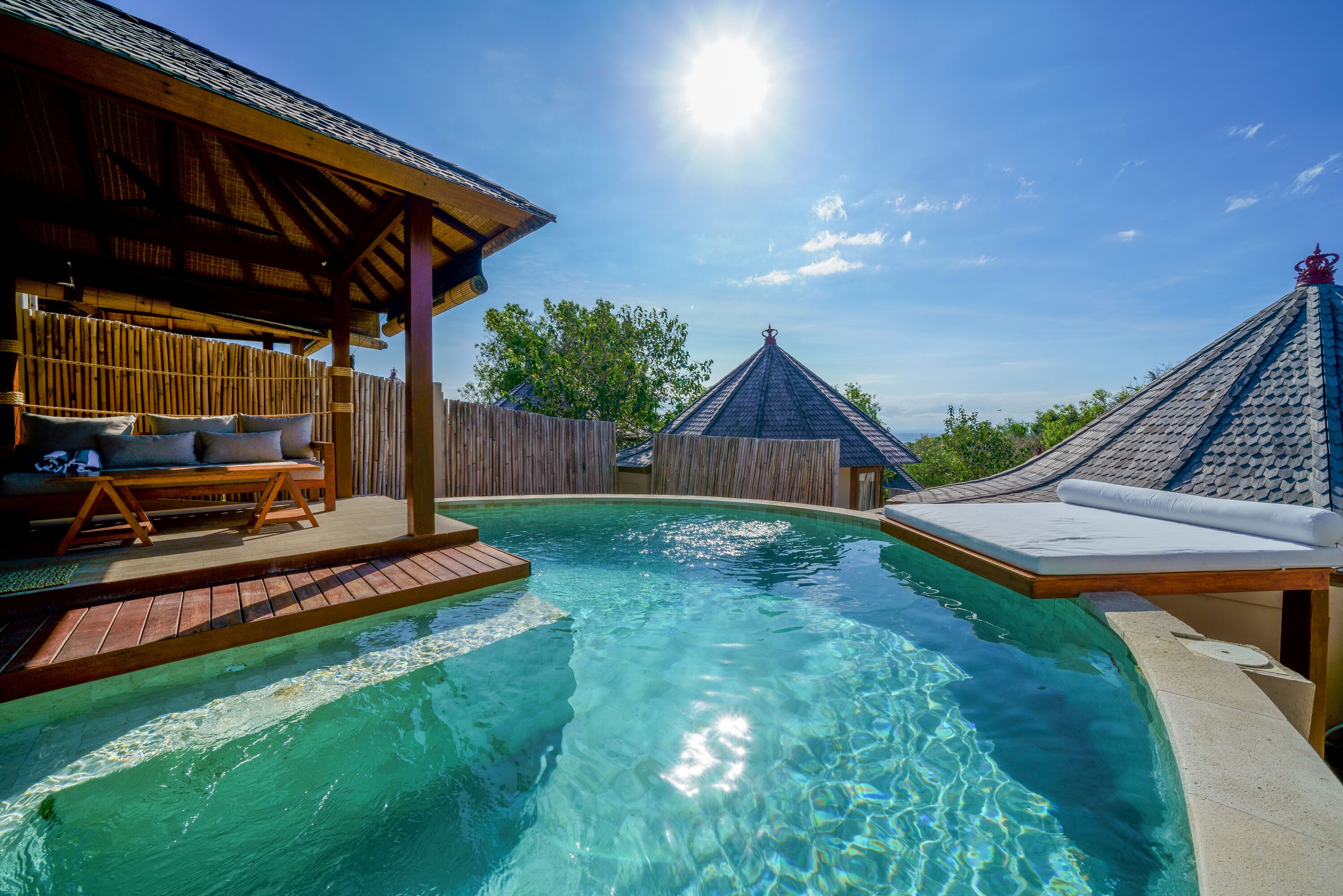 A swimming pool with wooden deck and lounge chairs, surrounded by wooden fences and a thatched-roof structure.