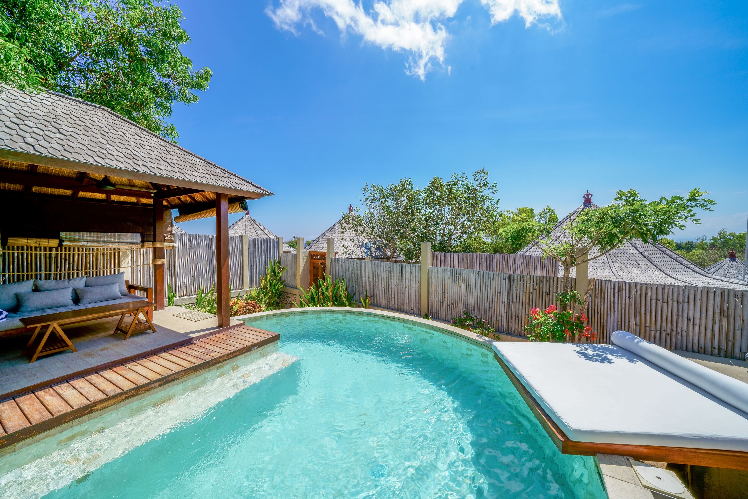 A backyard with a swimming pool, wooden deck, and a covered seating area with cushions.