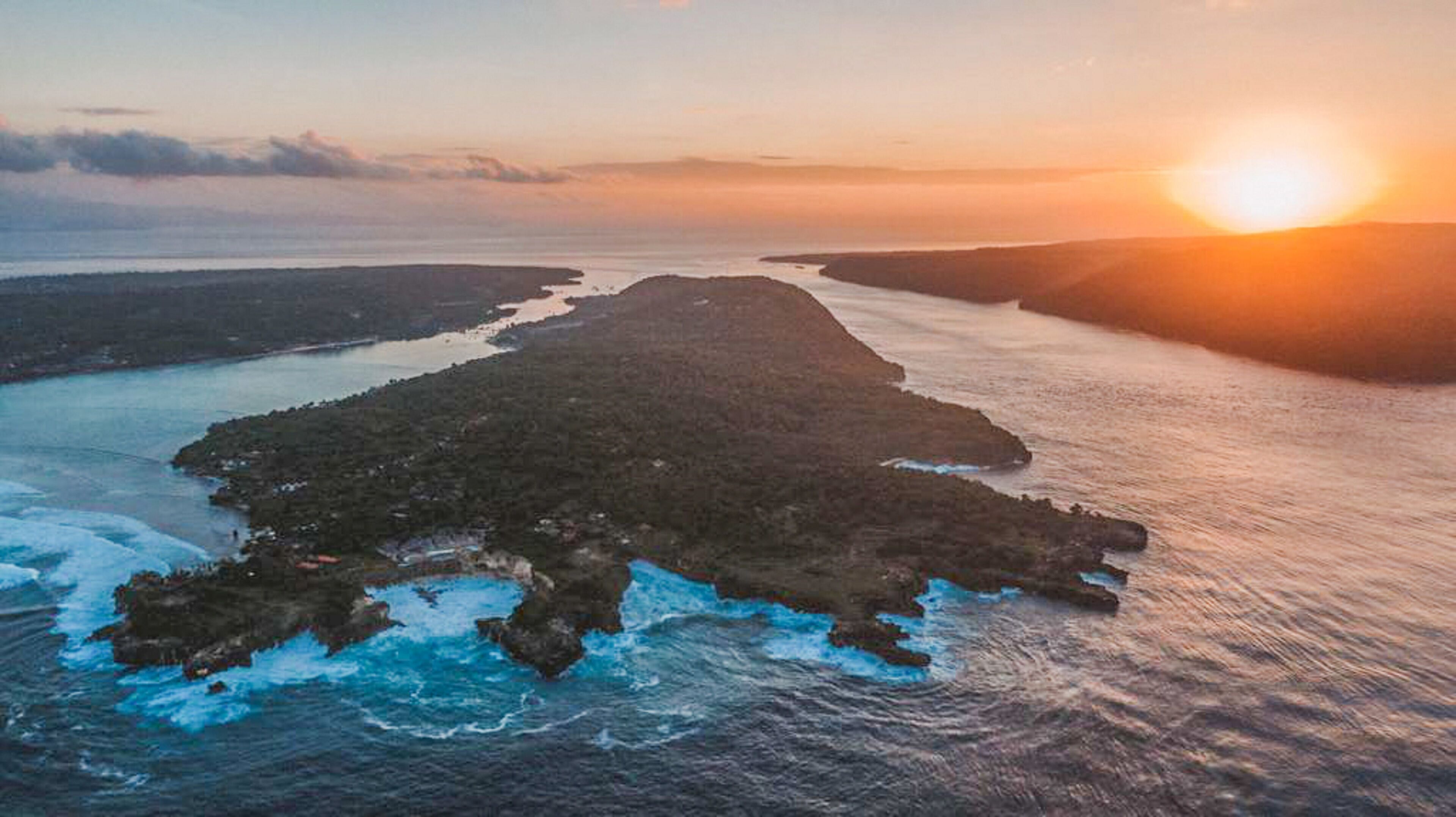 Vista aérea de un paisaje costero con una isla grande rodeada de agua, terreno rocoso y olas rompiendo contra la orilla.