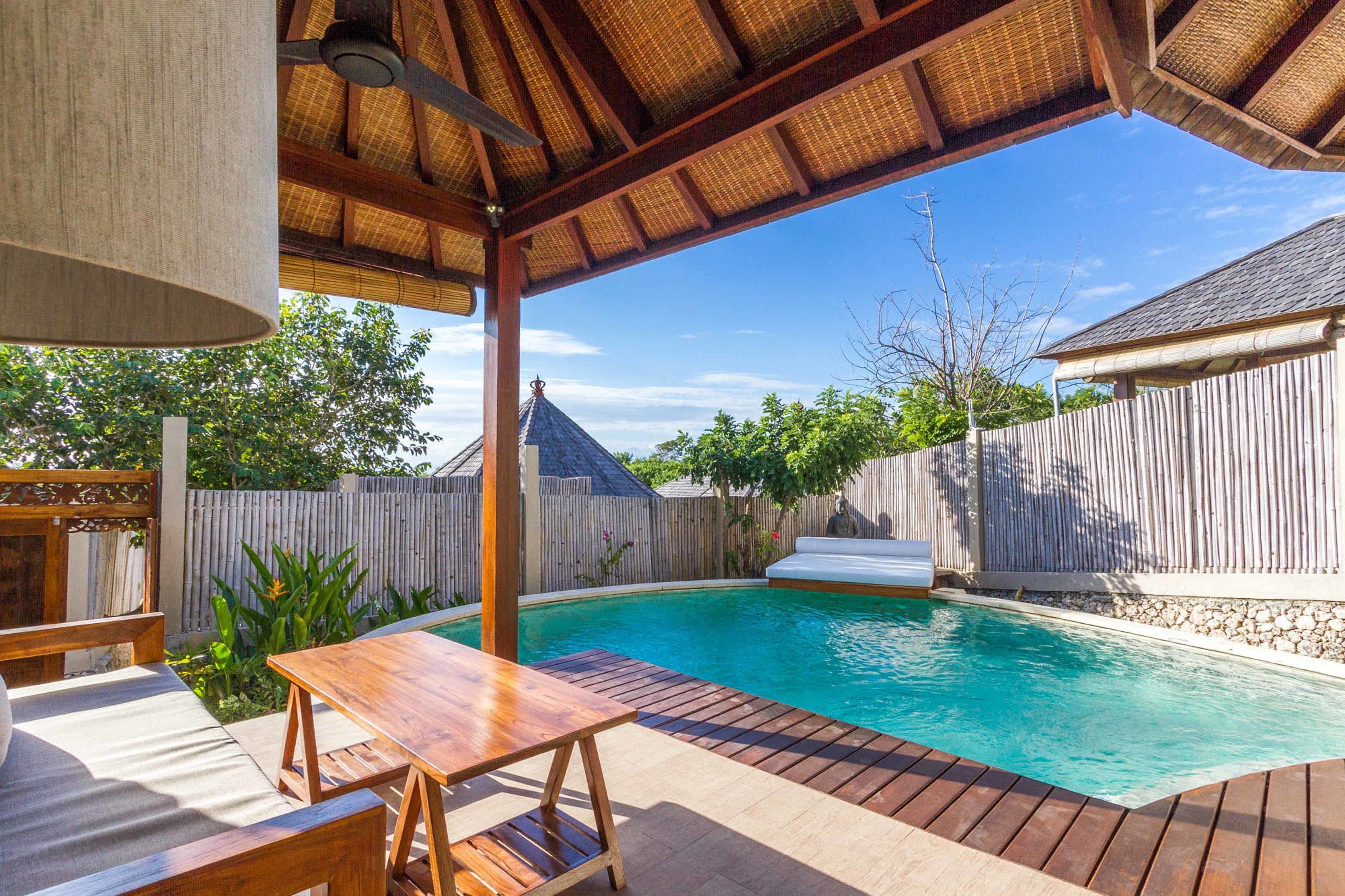 Una terraza de madera con piscina, un área de estar cubierta y una valla de madera al fondo.