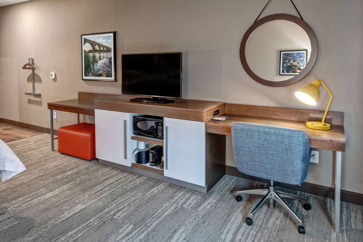 Habitación de hotel con un escritorio de madera, una televisión, un reposapiés rojo, una silla de oficina azul, una lámpara de escritorio amarilla y un cuadro en la pared.