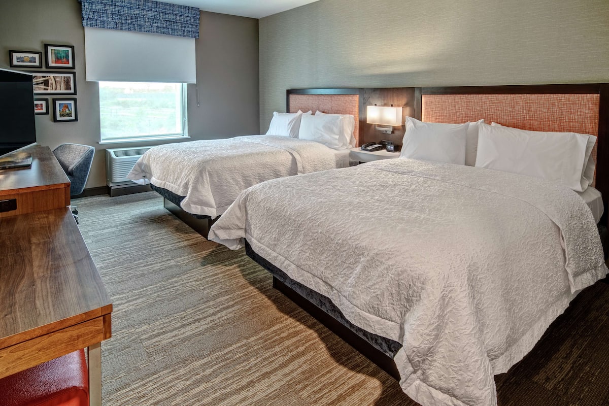 A hotel room with two beds, a desk, a chair, a TV, and a window with blinds.