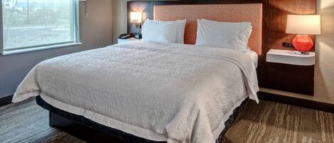 A hotel room with a large bed, a nightstand with a lamp, and a window with a view of the outdoors.
