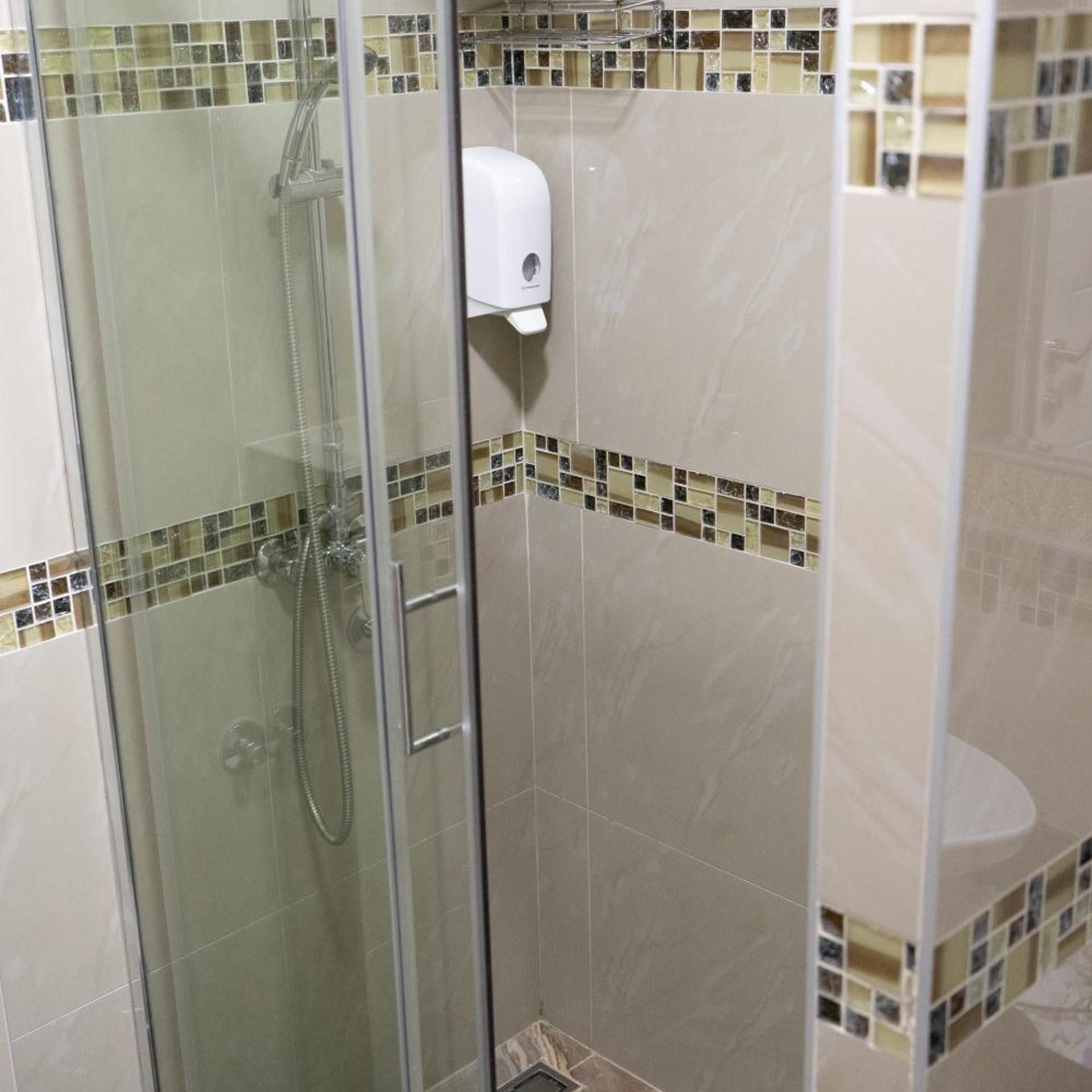 Une douche fermée par des parois en verre, dotée d’un pommeau de douche à main et d’un distributeur de savon fixé au mur.