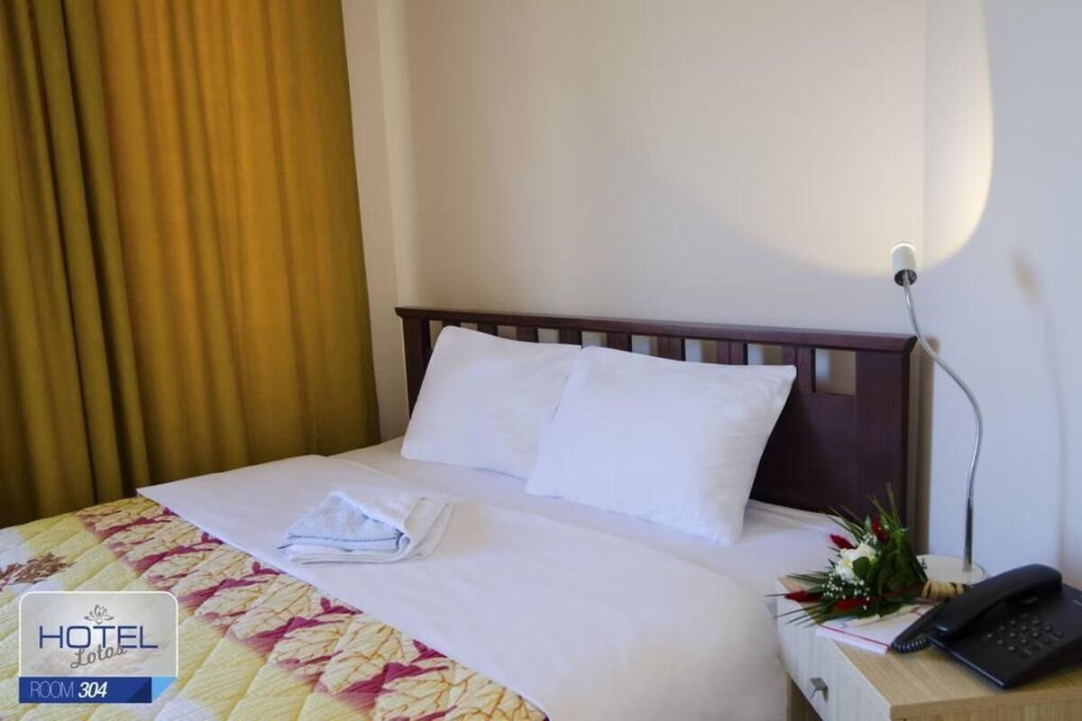 Ein Hotelzimmer mit einem ordentlich bezogenen Bett, einem Nachttischtelefon und einer Blumenkomposition auf dem Nachttisch.