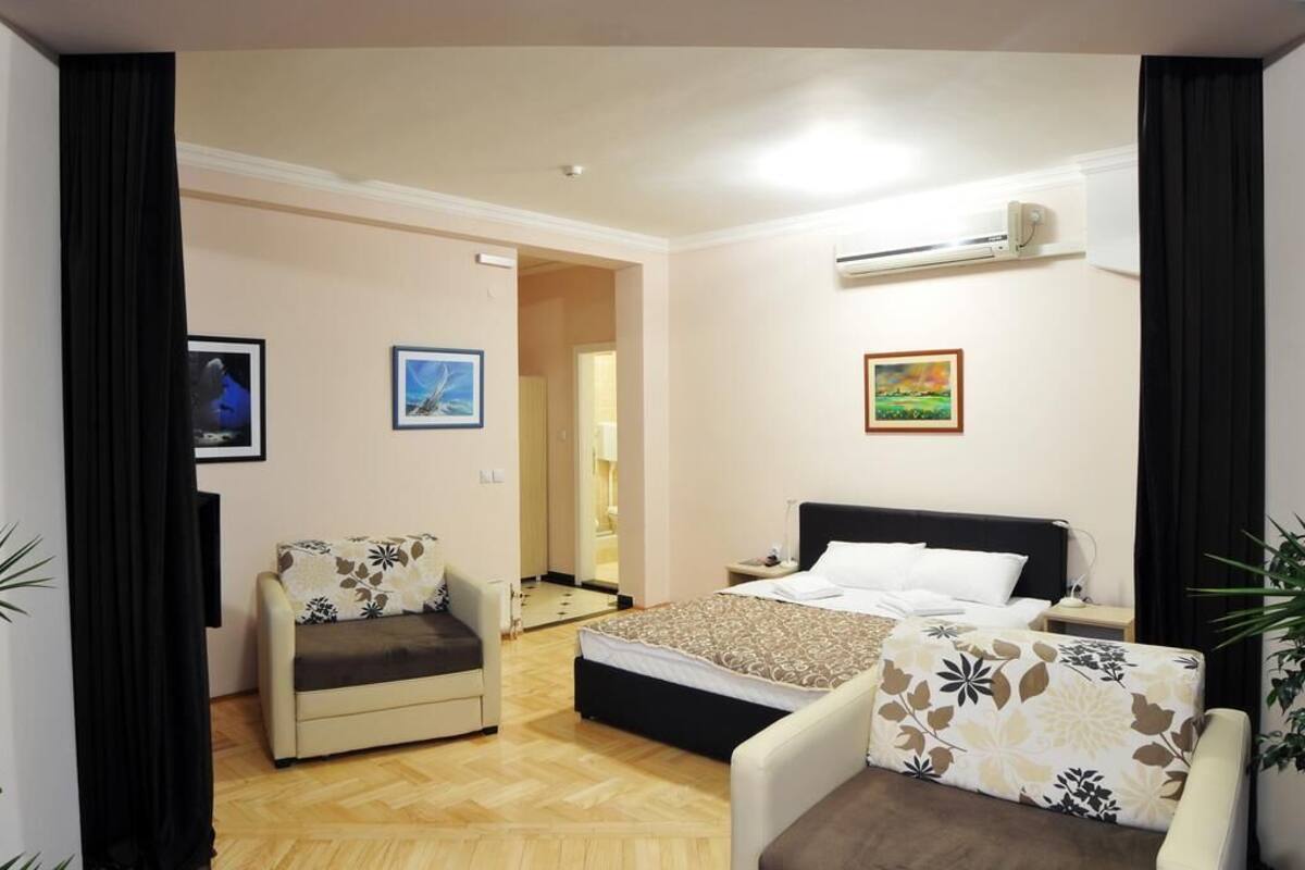 Ein Hotelzimmer mit einem Bett, einem Sofa, einem Sessel und einem kleinen Tisch. An den Wänden hängen zwei Gemälde und über dem Bett befindet sich eine Klimaanlage.