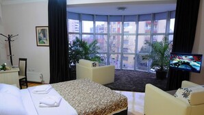 Une chambre d’hôtel avec un grand lit, deux fauteuils, une télévision et une vue sur les immeubles grâce à de grandes fenêtres.