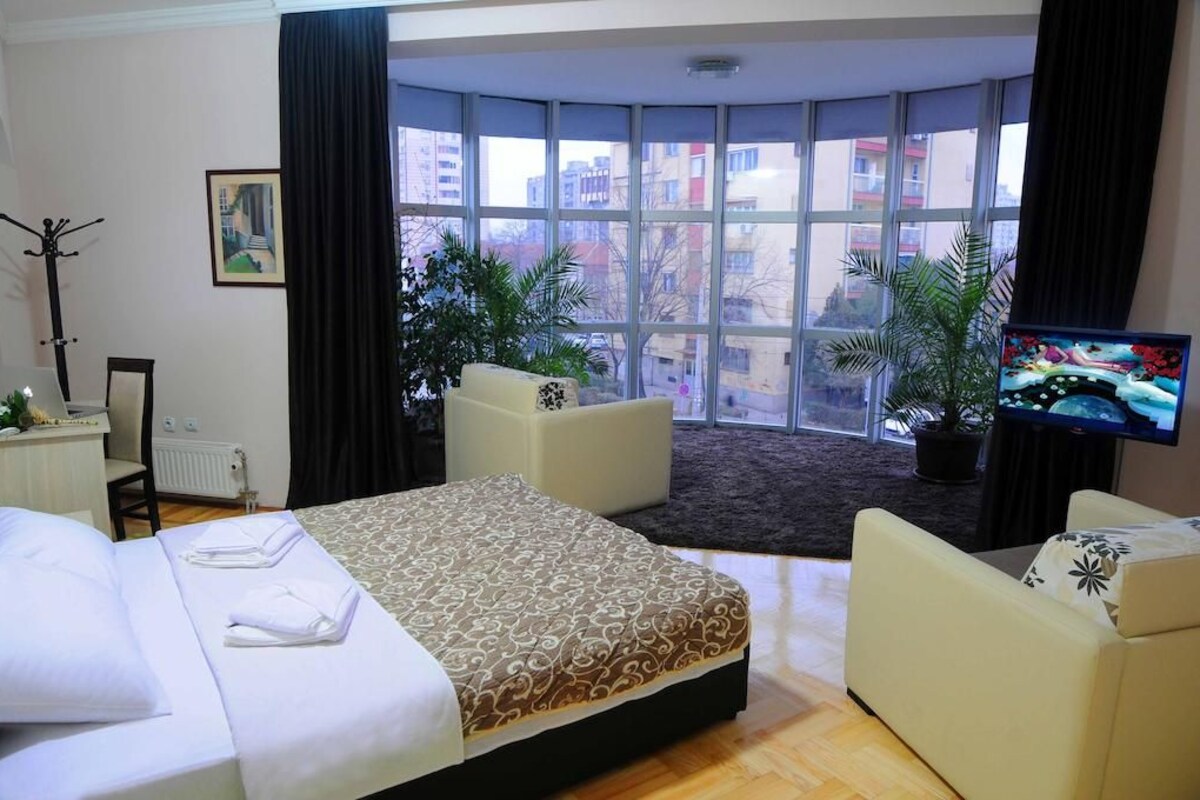 Une chambre d’hôtel avec un grand lit, deux fauteuils, une télévision et une vue sur la ville grâce à de grandes fenêtres.