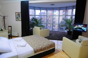 A hotel room with a large bed, two armchairs, a TV, and a view of the city through large windows. - Garni Hotel Lotos (Niš)
