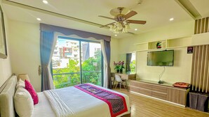 Una camera d'albergo moderna con un letto, una scrivania, una TV e un balcone con vista su edifici e vegetazione.