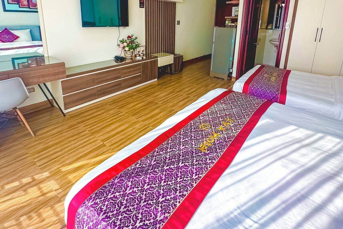 A modern hotel room with a wooden floor, a bed with a patterned bedspread, a desk with a chair, a TV mounted on the wall, and a ceiling fan.