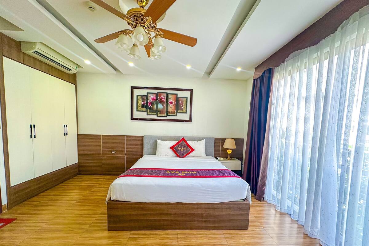 A hotel room with a large bed, wooden flooring, a ceiling fan, an air conditioning unit, a nightstand with a lamp, and a window with curtains.