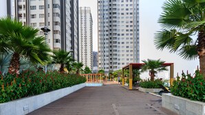 A wooden boardwalk with palm trees and red flowers, leading to a covered seating area with a view of tall buildings.