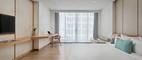 A modern hotel room with a large bed, a desk with a laptop, a TV, and a window with sheer curtains.