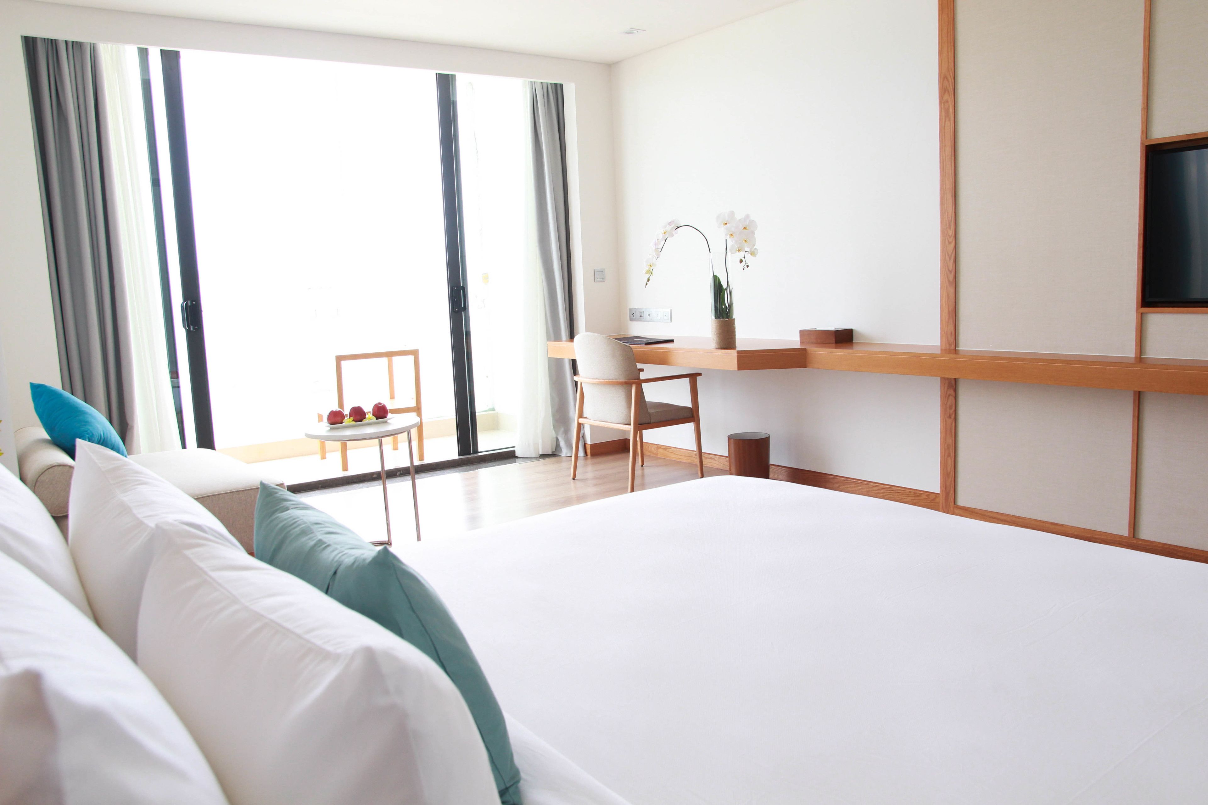 Habitación de hotel con una cama grande, un escritorio con silla, televisión y vistas al exterior a través de amplios ventanales.