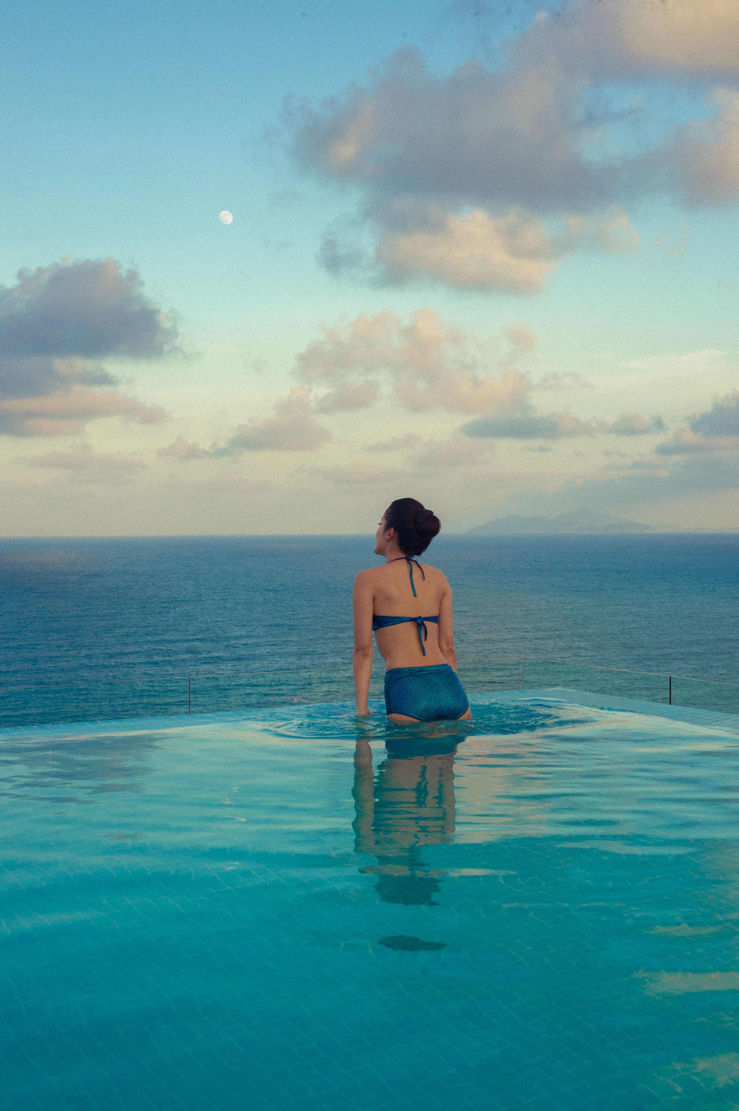 Una mujer con traje de baño azul está de pie en una piscina infinita azul cristalina, con vista al océano y una luna en el cielo.