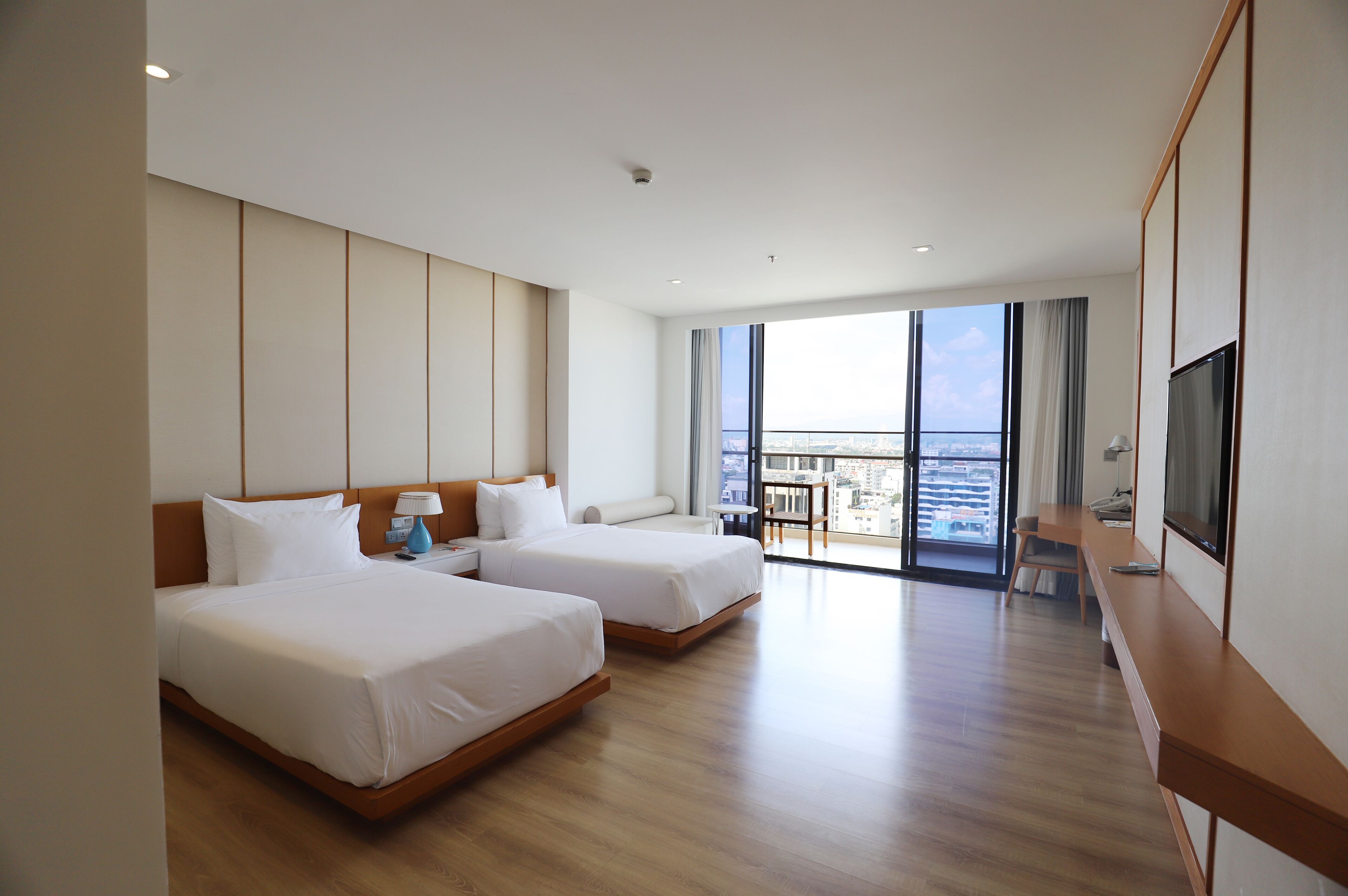 A hotel room with two beds, a desk, a TV, and a balcony with a city view.