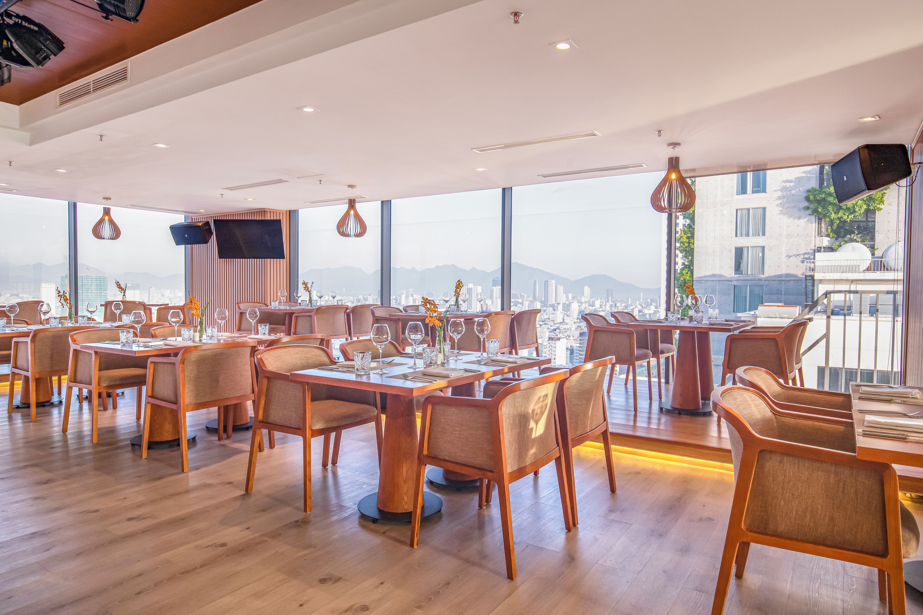 Un restaurante moderno con mesas y sillas de madera, amplias ventanas que ofrecen vistas de la ciudad e iluminación colgante.
