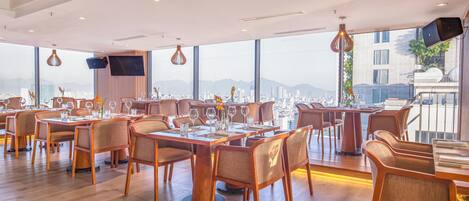 A modern restaurant with wooden tables and chairs, large windows offering city views, and pendant lighting.