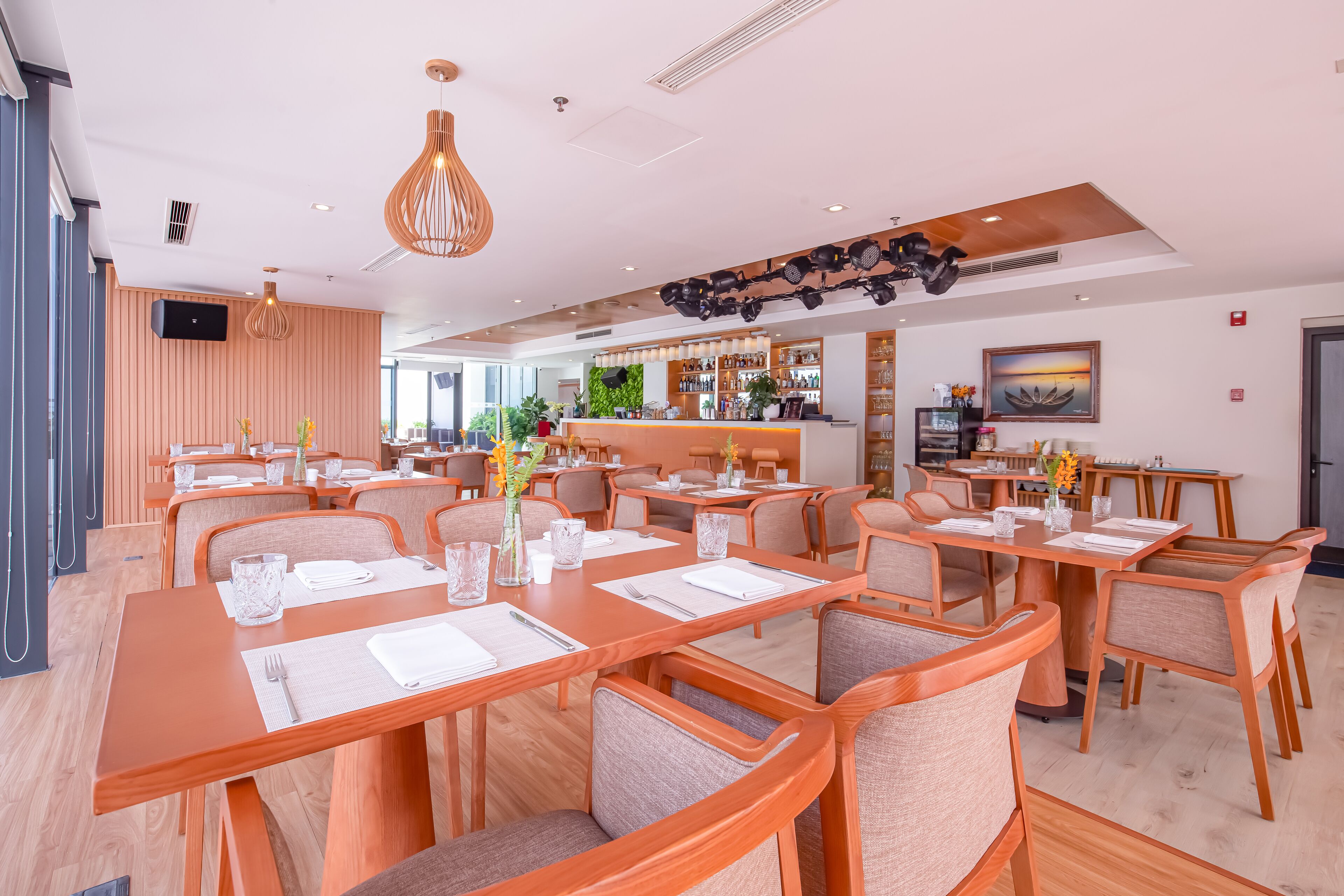 A modern restaurant with wooden tables and chairs, a bar area, and a large window providing natural light.