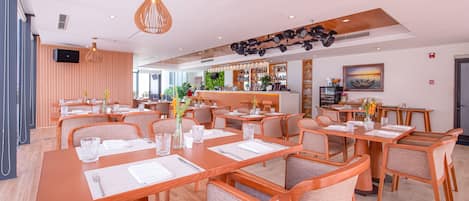 A modern restaurant with wooden tables and chairs, a bar area, and a large window providing natural light.