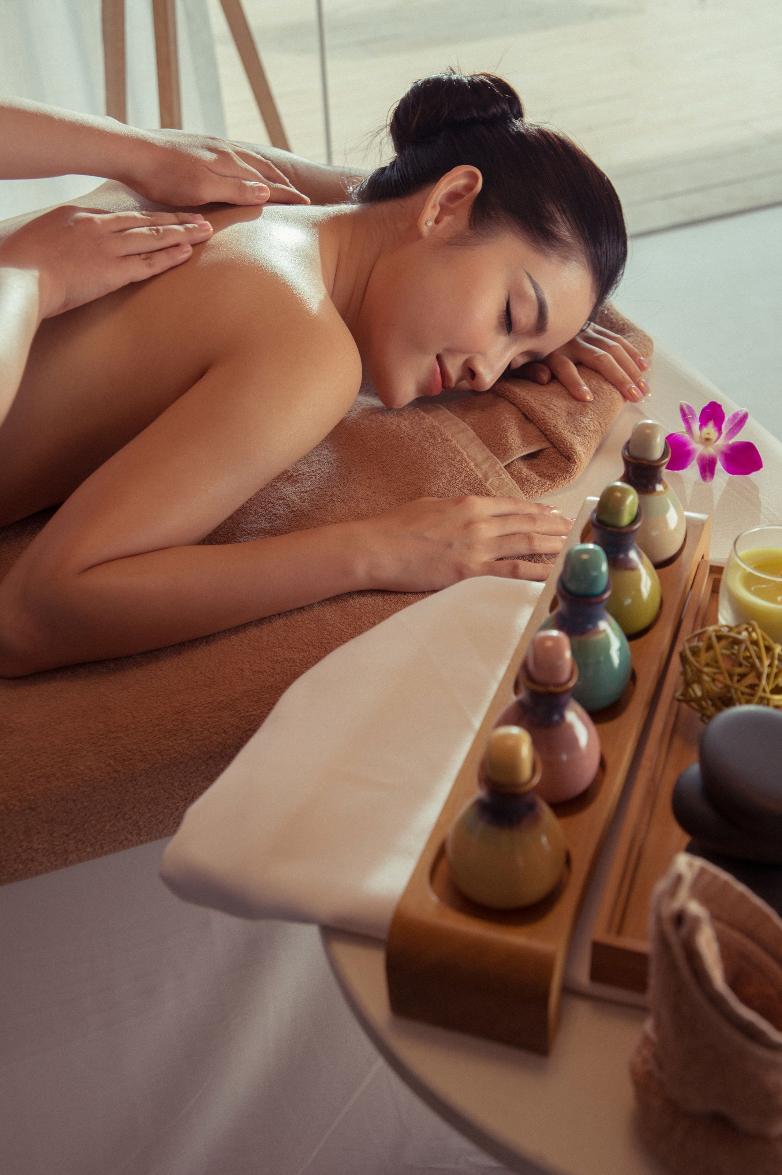 Una persona recibiendo un masaje en un spa, con una bandeja de aceites de masaje y una flor decorativa.