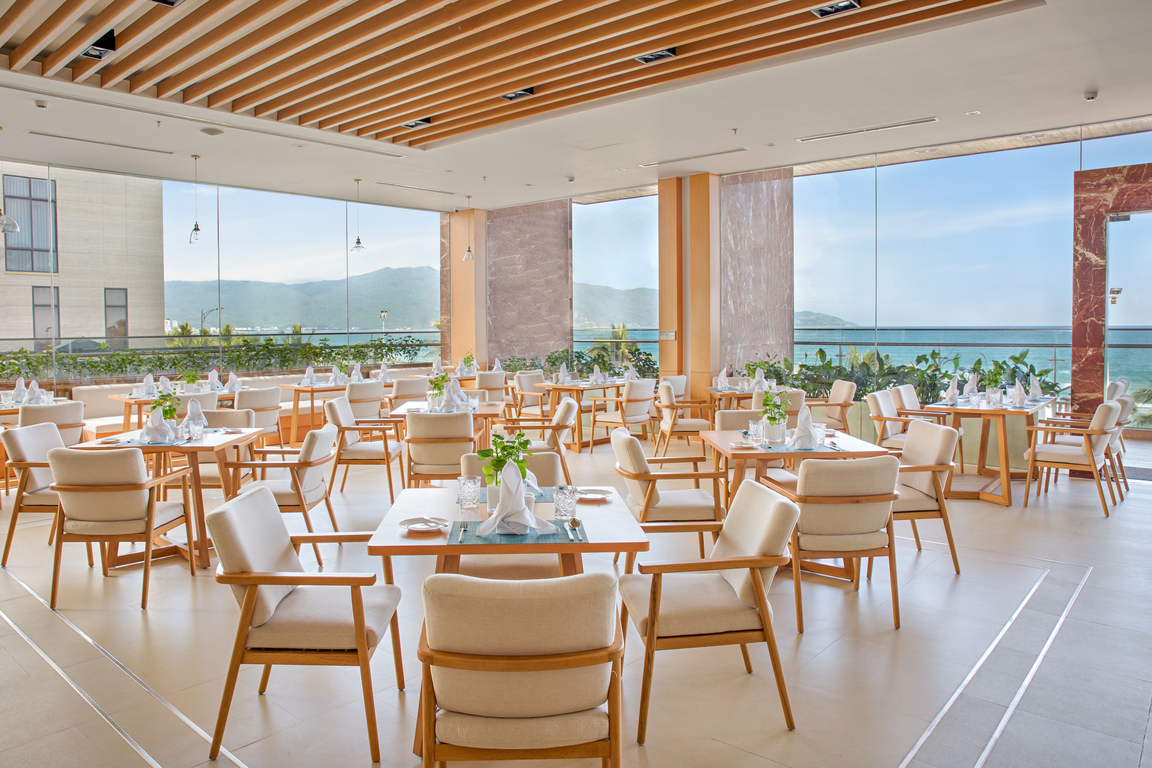 A spacious dining area with multiple tables and chairs, large windows offering a view of the sea and mountains.