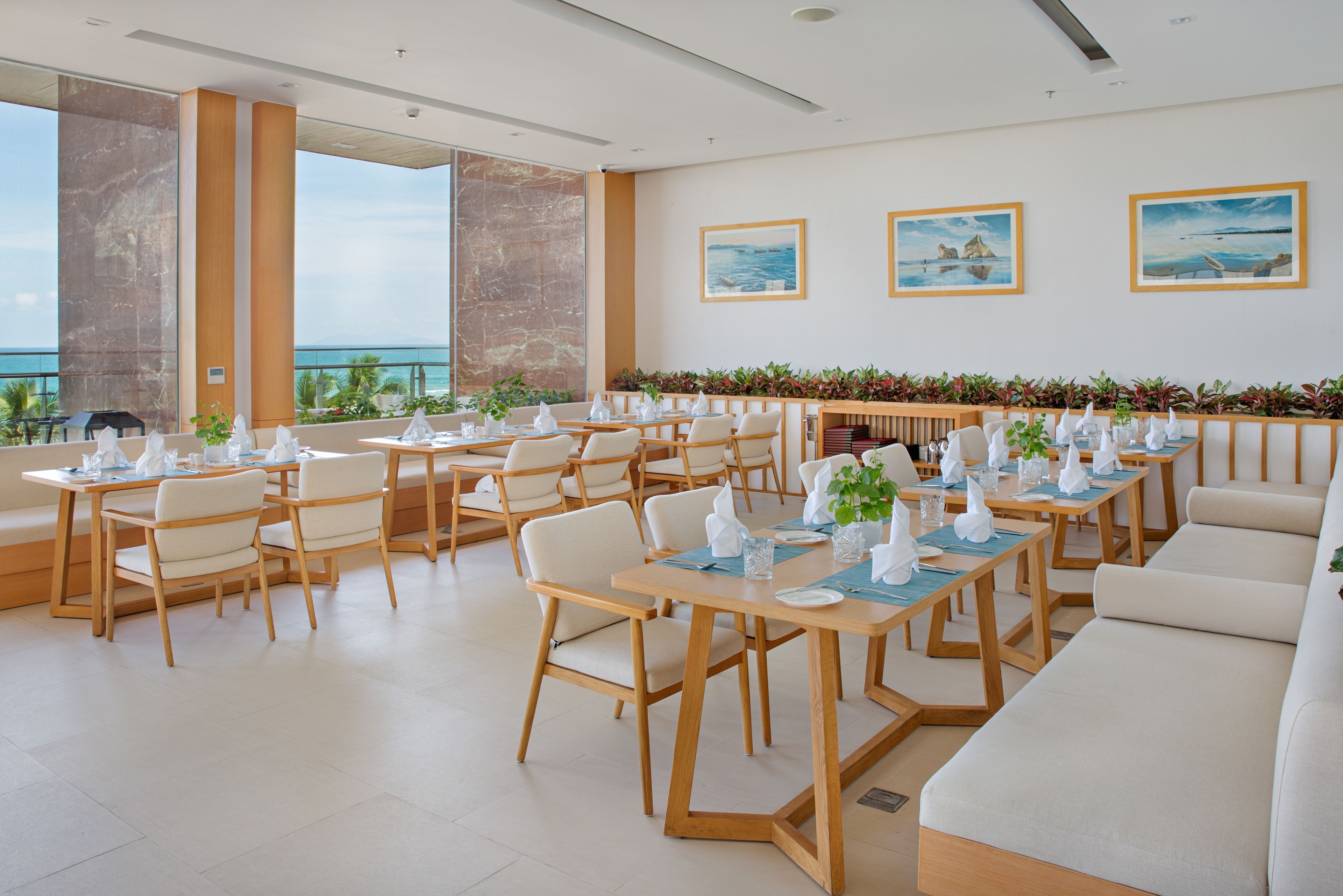 A spacious dining area with multiple tables and chairs, large windows offering a view of the ocean, and a balcony with plants.