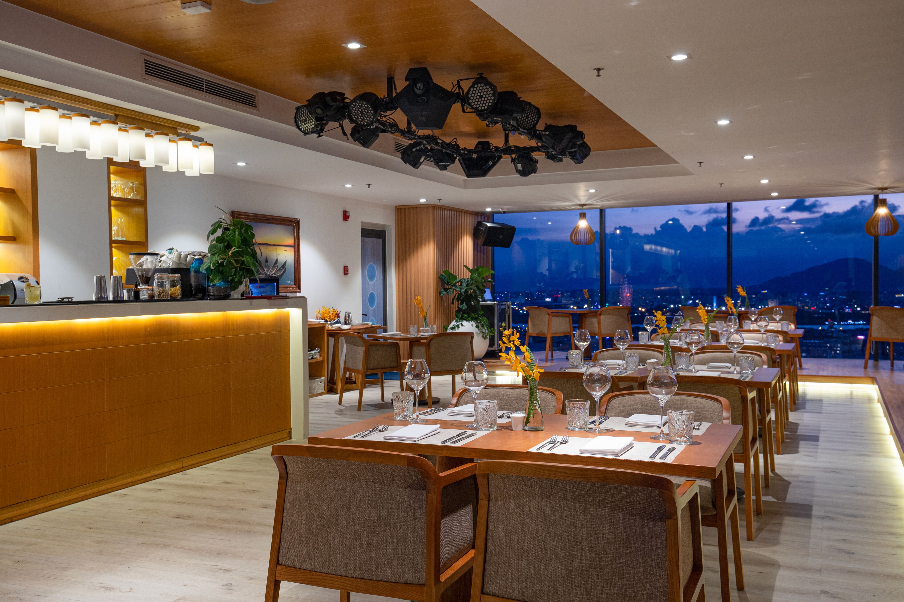 A restaurant with a long counter, multiple tables, and chairs, and a view of a cityscape at night.