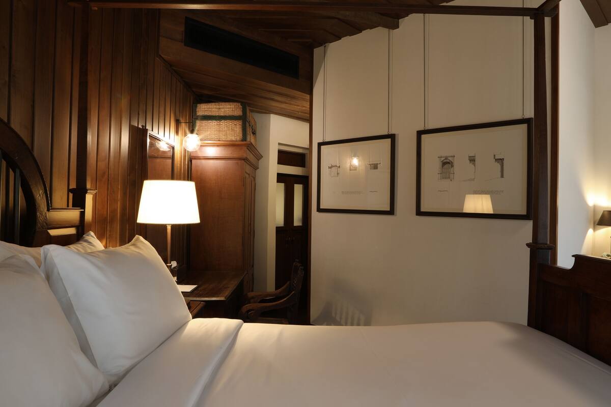 A hotel room with a wooden ceiling, a bed with white linens, a nightstand with a lamp, and two framed architectural drawings on the wall.