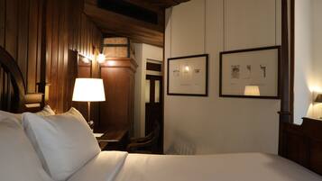 A hotel room with a wooden ceiling, a bed with white linens, a nightstand with a lamp, and two framed architectural drawings on the wall.