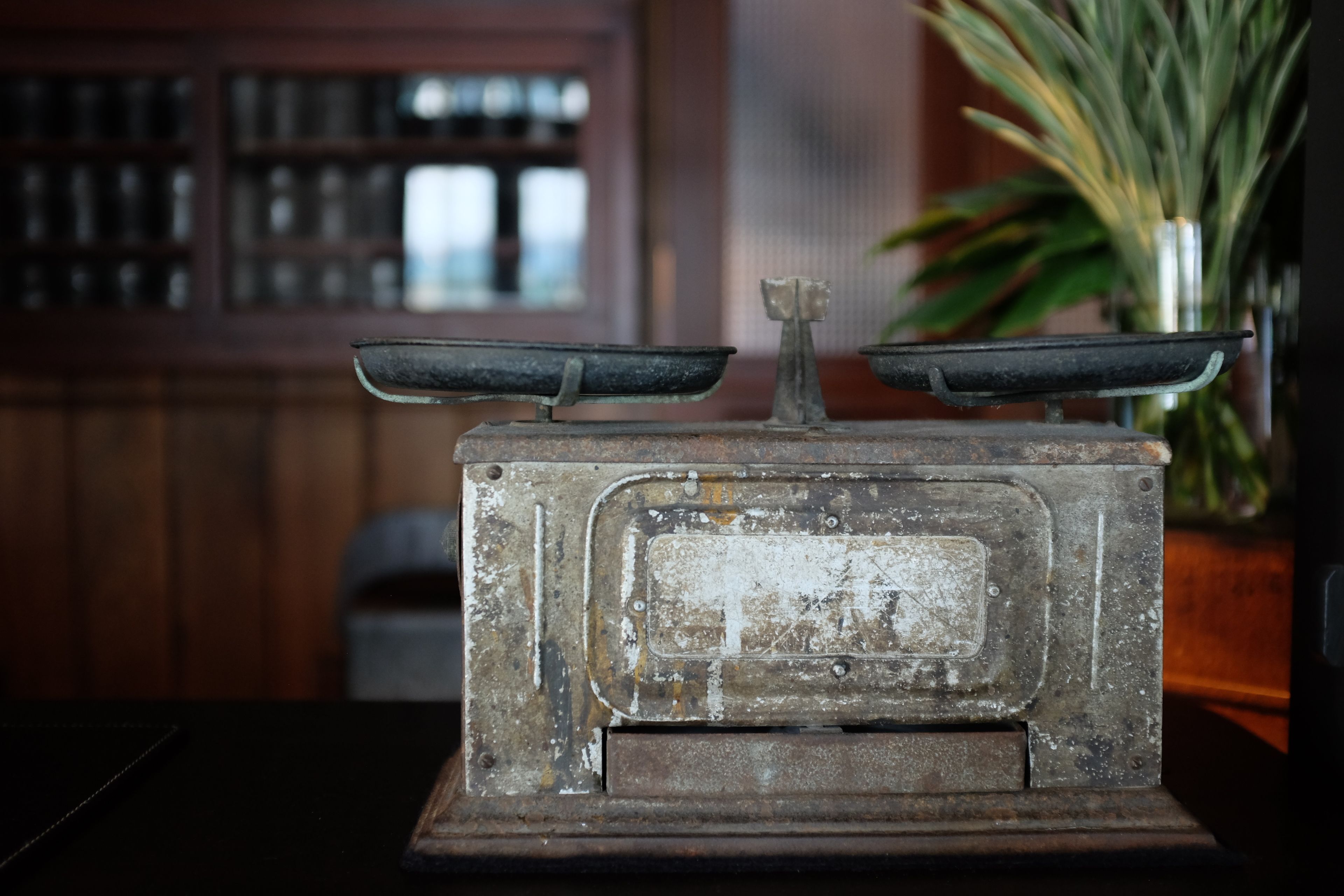 An old-fashioned scale with a wooden base, set against a blurred background with a plant and a window.