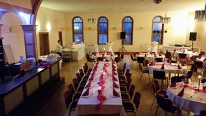 Salão de banquetes com longas mesas preparadas para um evento formal, com lustres e janelas em arco.