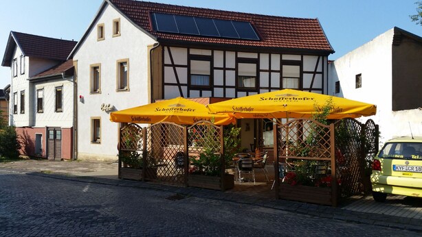 Um edifício tradicional europeu com fachada branca e estrutura de madeira escura. Na frente, áreas de estar ao ar livre com guarda-sóis amarelos com a inscrição "Schofferhofer".