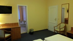 Quarto de hotel com cama, escrivaninha, cadeira, televisão, porta e banheiro visível através de uma porta aberta.