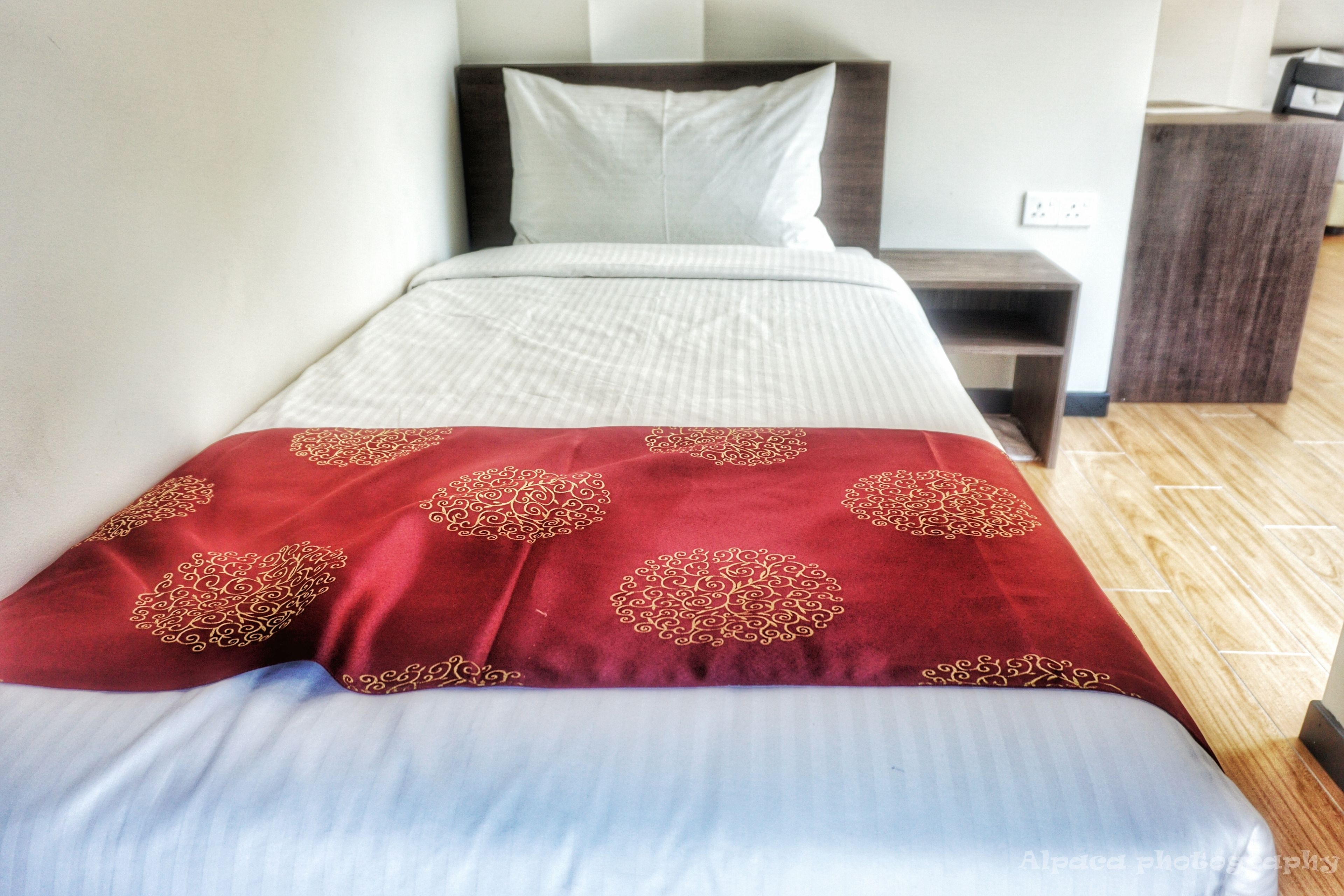 A neatly made bed with a white and red bedspread, a wooden nightstand with a drawer, and a wooden floor.